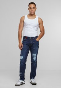 Mørkeblå denimjeans med slitte lapper, med kontraststing og røde detaljer på livet, kombinert med en hvit tanktopp.