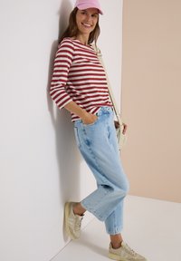 Femme souriante, portant une casquette rose, une chemise rayée rouge et blanche, un jean bleu clair, des baskets beige, appuyée contre un mur avec une main dans la poche.