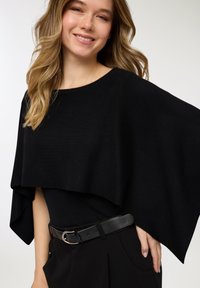Top nero oversize in stile poncho realizzato in tessuto morbido, con scollo ampio e maniche drappeggiate, abbinato a pantaloni neri a vita alta e cintura.
