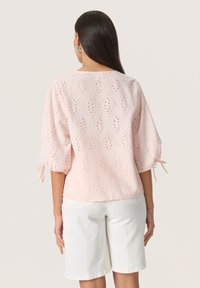 Blusa rosa chiaro con motivi intagliati a diamante, maniche ampie con legature e scollatura rotonda. Indossata con pantaloni corti bianchi, che mostrano una vestibilità rilassata.