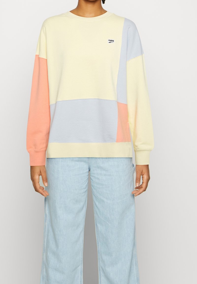 Personne portant un sweat-shirt color-block avec des panneaux jaune pastel, bleu et pêche, associé à un jean bleu clair sur un fond uni.