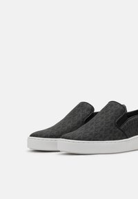 Chaussures à enfiler avec une tige en tissu noir texturé, des panneaux latéraux élastiques et une semelle en caoutchouc blanche, présentant un motif subtil dans le matériau.