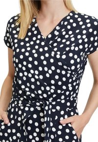 Femme portant une robe portefeuille bleu marine à pois blancs, manches courtes et ceinture en tissu nouée à la taille.