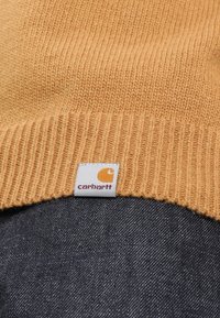 Brązowa dzianinowa czapka beanie o prążkowanej fakturze, z białą metką z pomarańczowym logo, umieszczona na ciemnym dżinsowym materiale.