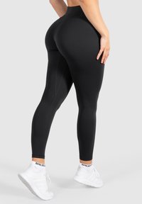 Leggings neri a vita alta realizzati in tessuto elastico, dotati di un design senza cuciture, texture liscia e gambe affusolate. Indossati con scarpe sportive bianche.
