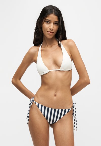 Femme aux longs cheveux foncés portant un haut de bikini blanc à col halter et un bas de bikini rayé bleu marine et blanc avec des liens sur les côtés.
