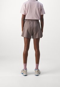 Camisa de algodón rosa de manga corta combinada con pantalones cortos marrones. El modelo lleva zapatillas ligeras con acentos azules y calcetines rosas, mostrado desde la parte de atrás.