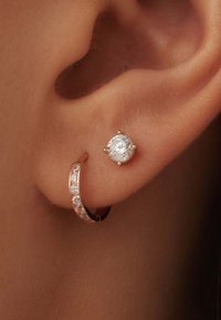 Boucle d'oreille en or rose ornée d'une rangée de petits cristaux, associée à une boucle d'oreille à clou avec une pierre ronde et claire, sur un fond de teinte de peau.