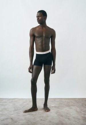 Νεαρός μαύρος άνδρας που στέκεται, φοράει μαύρα σλιπ μπόξερ με λευκή και πορτοκαλί μέση, κοιτάζει αριστερά μπροστά σε ένα απλό φωτεινό φόντο.