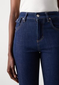 Mørke denimjeans med høy midje, intrikate sømmer og detaljer i sølvmetall. Glatt tekstur med svak glans.