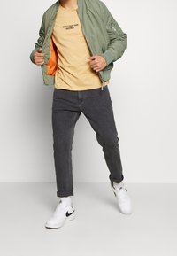 Grüne Bomberjacke mit orangefarbenem Innenfutter, beiges T-Shirt mit Aufdruck, dunkelgraue Jeans und weiße Sneaker mit schwarzem Akzent.
