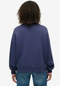 Navy blue oversize sweatshirt z miękkiego materiału, z opuszczonymi ramionami i okrągłym dekoltem, w zestawieniu z jasnoniebieskimi dżinsami i różową warstwową koszulą.