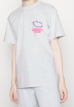 Osoba ubrana w jasnoszary T-shirt z fioletową buźką i różowym napisem "Carhartt WIP" na lewej piersi, w połączeniu z jasnoniebieskimi dżinsami.