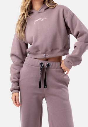 Kobieta w mauve przyciętej bluzie z kapturem z napisem „Just Hype” i dopasowanych spodniach dresowych ze sznurkiem, ręka na biodrze, widoczne jasne falowane włosy.