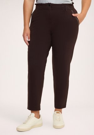Donna che indossa pantaloni marrone scuro a trombetta con tasche laterali abbinati a sneakers bianche, in piedi davanti a uno sfondo semplice.