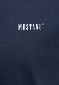 Tmavomodrá látka s bielym veľkými tlačenými písmenami "MUSTANG" v strede.