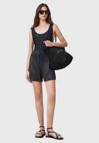 Top nero senza maniche con ripetizione del testo "ALLSAINTS", pantaloni corti in rete nera e sandali neri con cinturini. Il modello porta una grande borsa nera con logo.