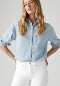 Camisa de mezclilla azul claro con botones y mangas largas, arremangadas en los puños, que presenta un bolsillo en el pecho y un pequeño logo de Levi's. Llevada con jeans blancos.