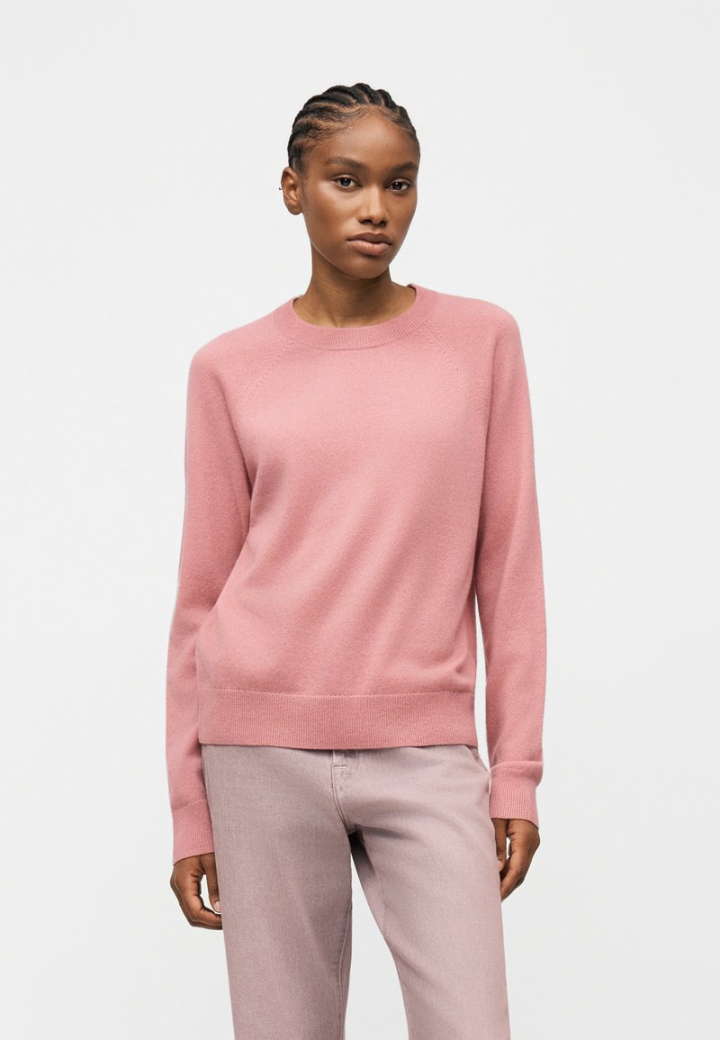 Femme aux cheveux tressés portant un pull ras du cou rose clair et un pantalon rose pâle, debout devant un fond uni clair.