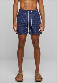 Shorts de bain navy avec des motifs nautiques blancs, taille élastique, cordon de serrage et poches latérales. Portés avec des sandales brunes.