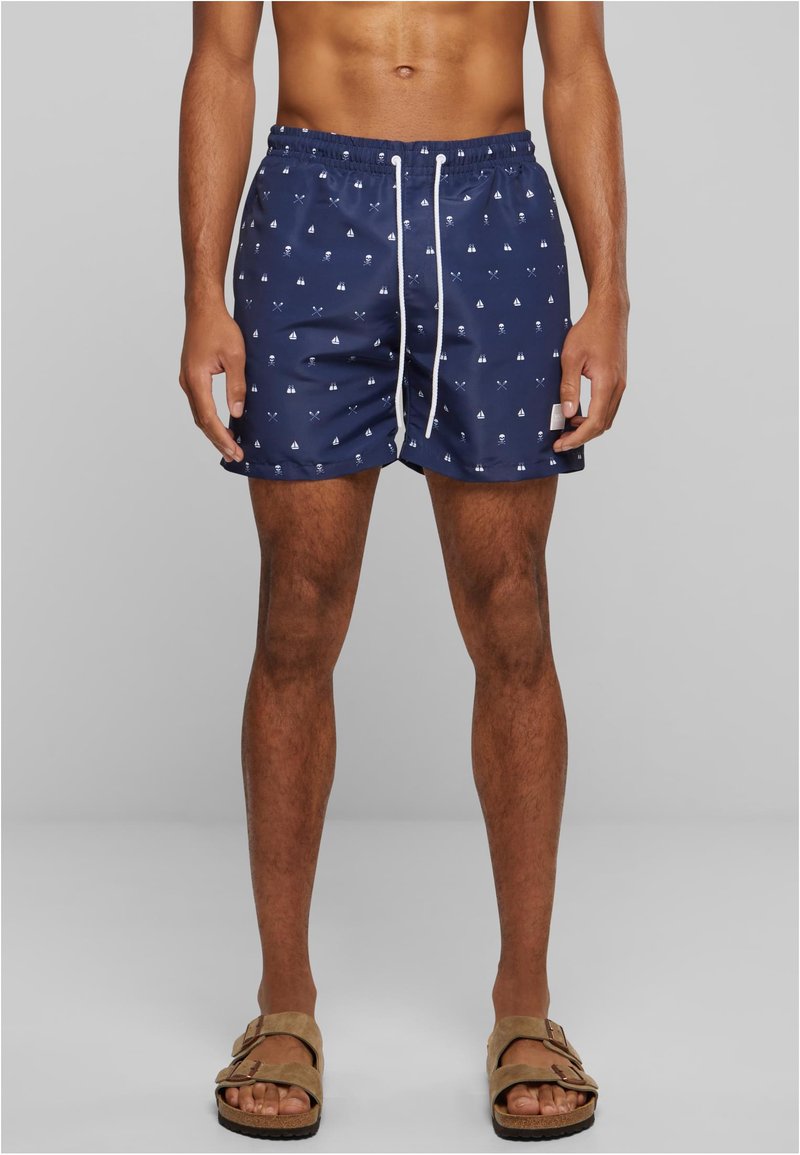 Shorts de bain navy avec des motifs nautiques blancs, taille élastique, cordon de serrage et poches latérales. Portés avec des sandales brunes.