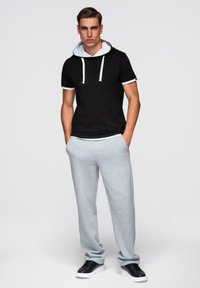 Mladý muž stojící s rukama v kapsách, oblečený v černé mikině s krátkým rukávem, šedých teplákách a černých teniskách na jednobarevném pozadí.