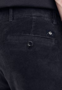 Pantalons en velours côtelé bleu marine foncé avec un biject au tissu texturé, dotés d'une poche arrière unique avec une étiquette noire et d'une fermeture à bouton.