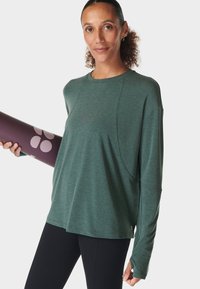 Vrouw met krullend haar in een groene lange mouw top en zwarte leggings, die een paarse yogamat met roze cirkels vasthoudt, staand tegen een witte achtergrond.