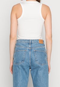 Top blanco sin mangas con un diseño ajustado, combinado con unos jeans de mezclilla de corte alto de color azul claro que tienen un parche de cuero marrón y dos bolsillos traseros.