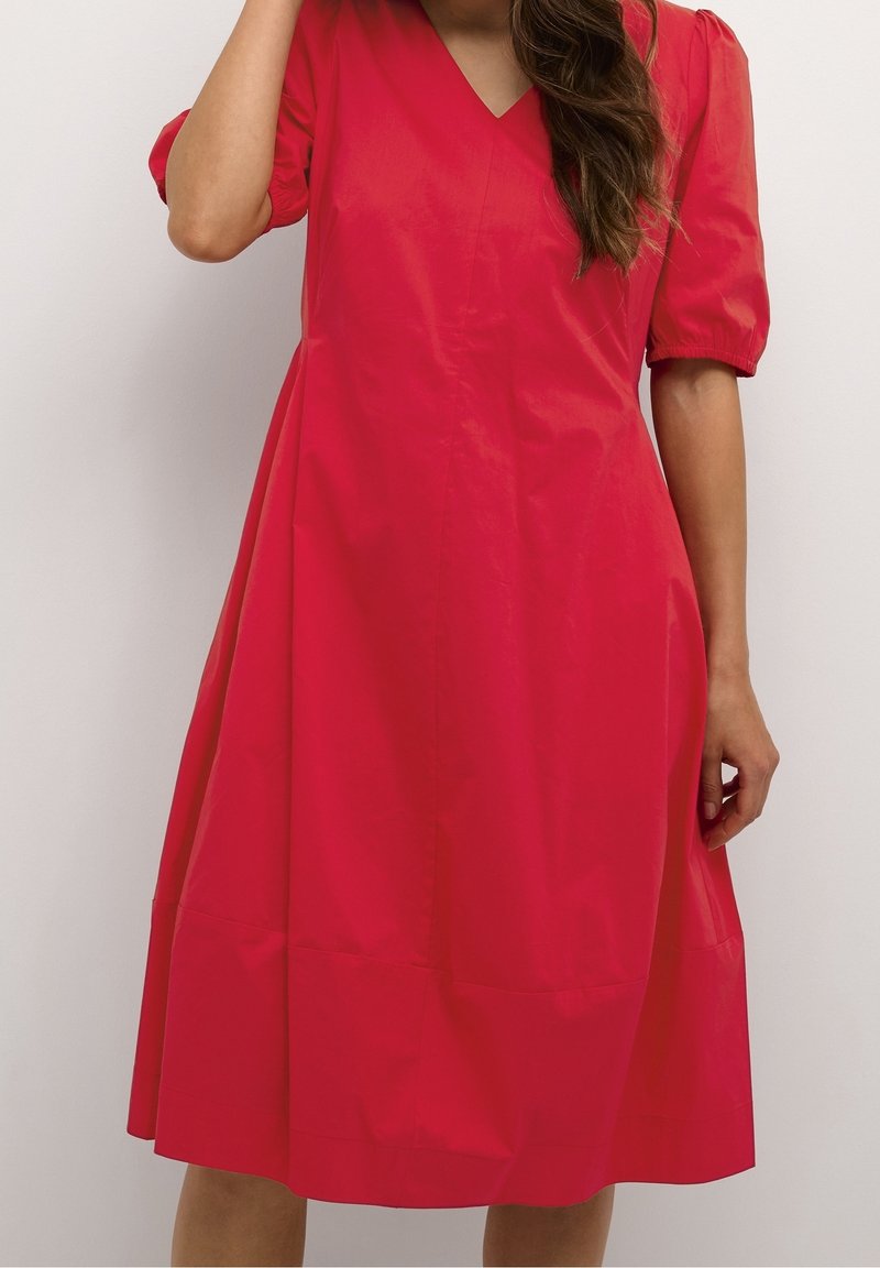 Nainen, joka on pukeutunut kirkkaan punaiseen polvipituiseen mekkoon, jossa on lyhyet puffihihat ja V-pääntie, seisoo vaaleaa yksinkertaista taustaa vasten.