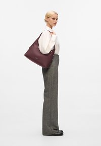 Borsa a spalla in pelle bordeaux con hardware argento, texture liscia e forma oversized. Il modello indossa pantaloni grigi e una camicia bianca.