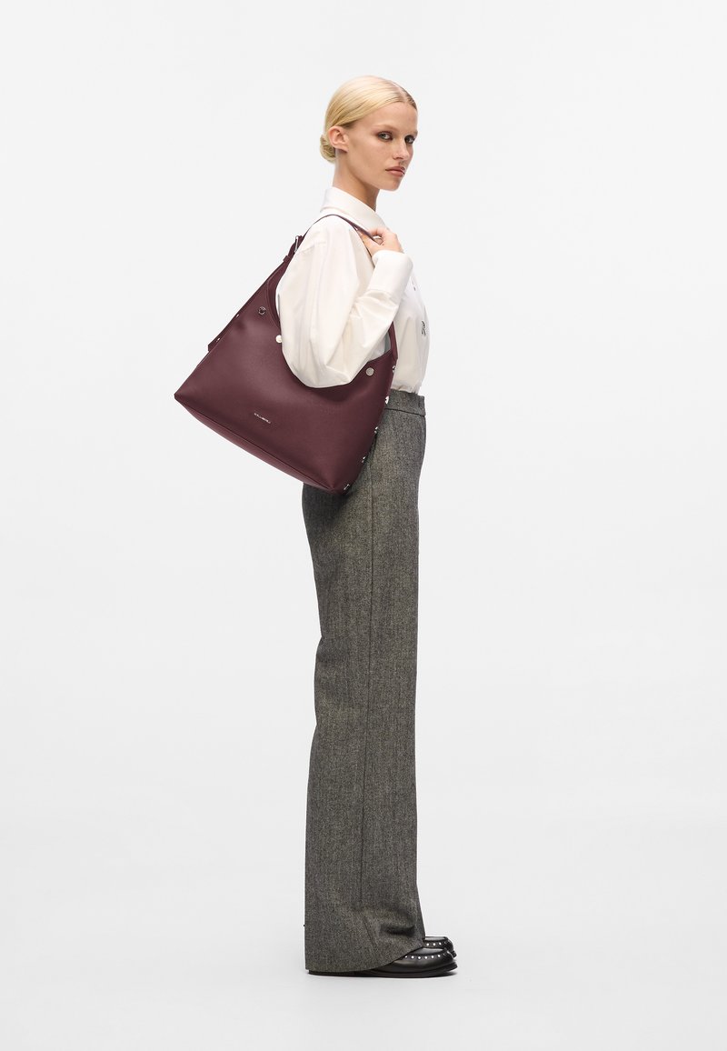 Borsa a spalla in pelle bordeaux con hardware argento, texture liscia e forma oversized. Il modello indossa pantaloni grigi e una camicia bianca.