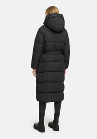 Black long puffer coat with a hood, quilted design, belted waist, and thick padding. Worn with black fitted pants and boots.