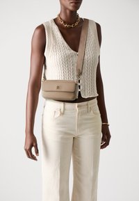 Sac en cuir beige en bandoulière de forme rectangulaire, avec des accessoires en or et un logo en relief. Porté avec un haut en tricot crème à torsades et un jean clair.