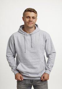 Giovane in un hoodie grigio chiaro con le mani nelle tasche, in piedi contro uno sfondo bianco semplice, che sorride leggermente.