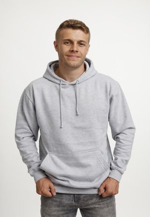 Jonge man in een lichtgrijze hoodie met zijn handen in zijn zakken, staand tegen een eenvoudige witte achtergrond, met een lichte glimlach.