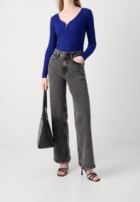 Haut bleu à manches longues côtelé avec une fermeture éclair, associé à un jean noir taille haute à jambes larges. Un sac à main noir et des talons noirs à lanières complètent le look.