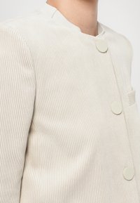Giacca in corduroy beige chiaro con collo alto, dotata di bottoni rotondi e piatti e una tasca frontale singola. Sottile texture a coste.