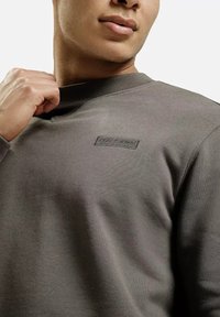 Szary sweter z teksturowanej tkaniny, okrągłym dekoltem i małą naszywką z logo na piersi. Gładka powierzchnia i dopasowany krój.