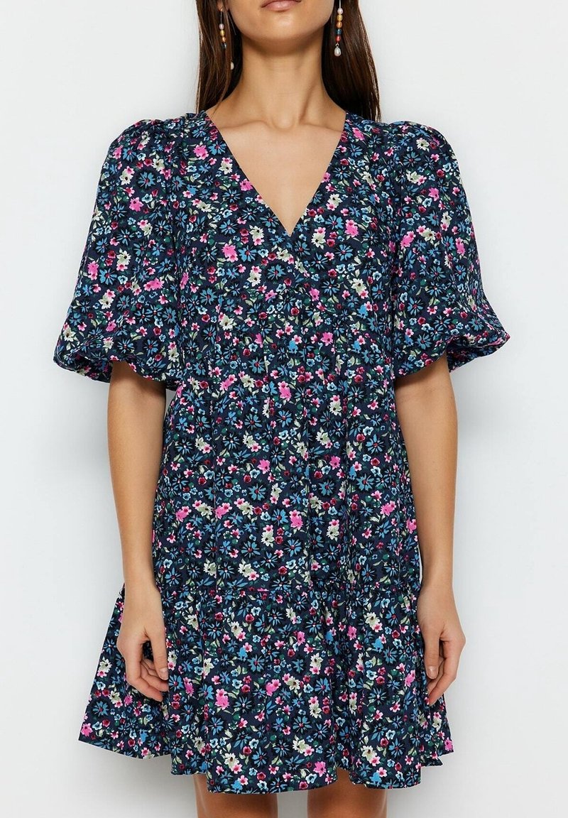 Frau trägt ein marineblaues Blumenkleid mit rosa, weißen und blauen Blumen, mit Puffärmeln und V-Ausschnitt vor einfachem Hintergrund.
