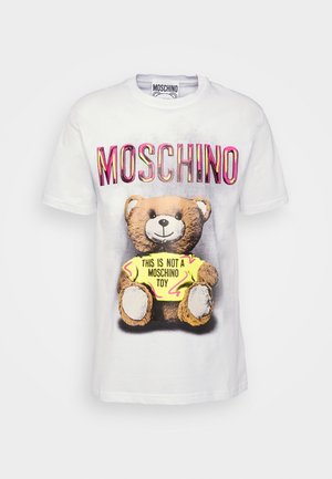 Henkilö, jolla on valkoinen Moschino-t-paita, jossa on nallekarhukuva ja mustat housut, joissa on keltaisia ja sinisiä geometristä kuvioita.