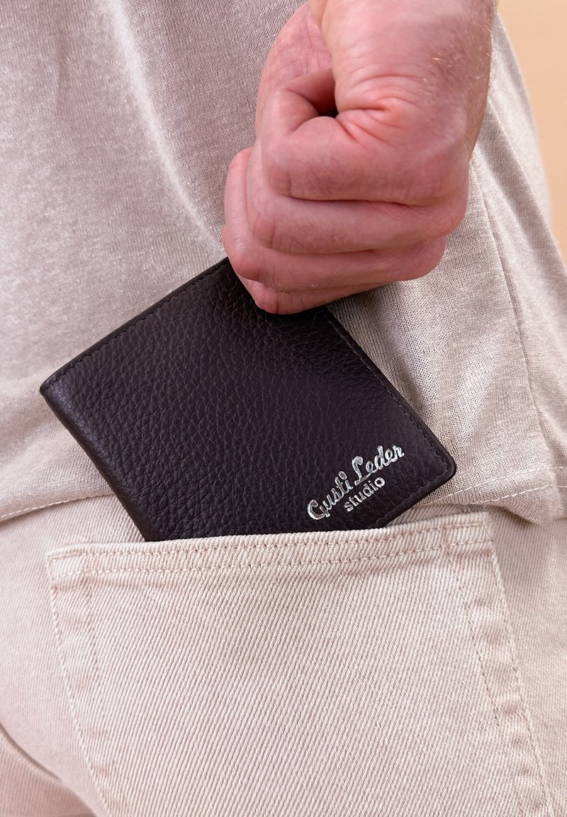 Tmavě hnědá kožená peněženka částečně zasunutá do béžové kapsy, s texturovaným povrchem a stříbrným logem v pravém dolním rohu.