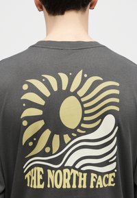 Άτομο που φοράει σκουρόγκρι μπλουζάκι με κίτρινο ήλιο και λευκό σχέδιο κύματος πάνω από το κείμενο «THE NORTH FACE» στην πλάτη.