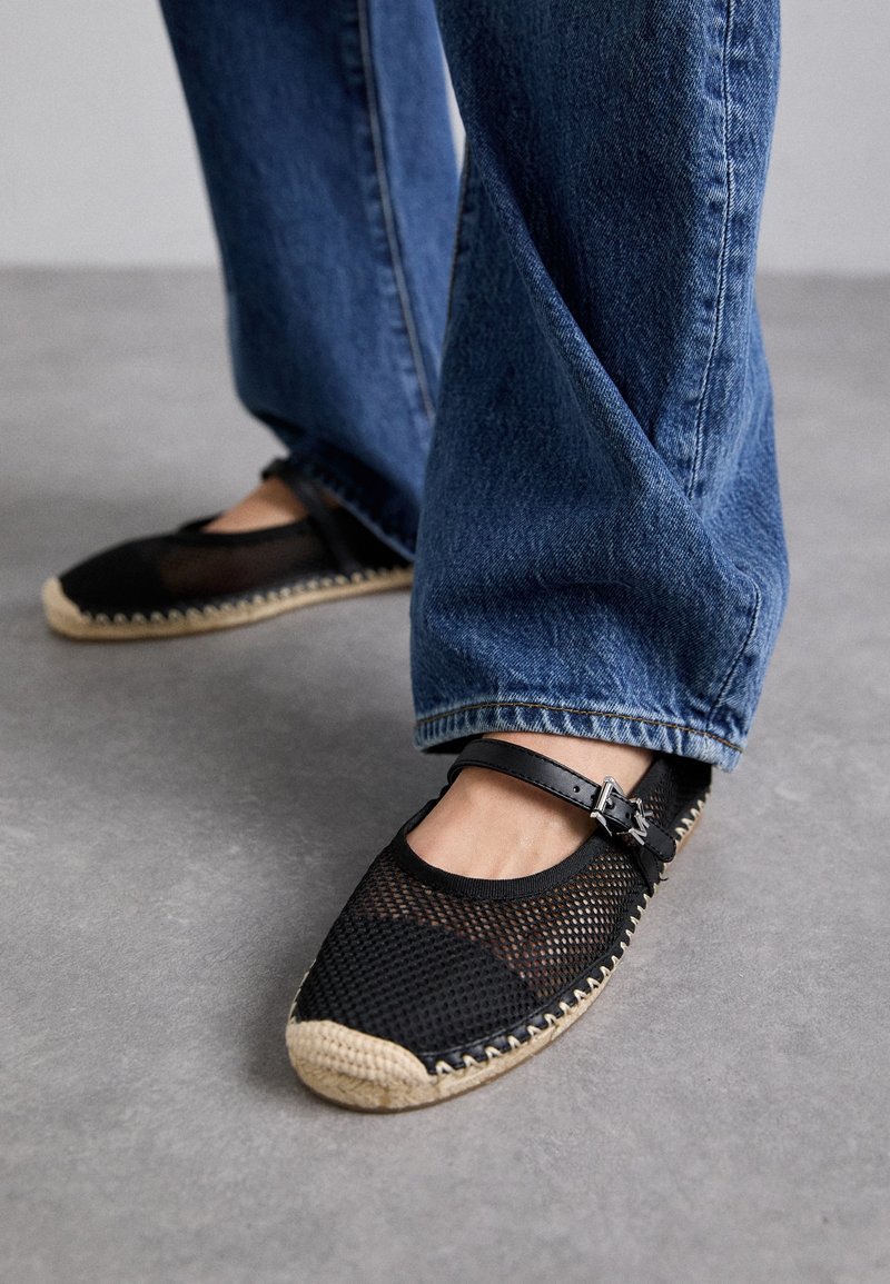 Pieds portant des espadrilles Mary Jane en mesh noir avec des semelles tissées beiges, associées à un jean bleu ample sur un sol gris.