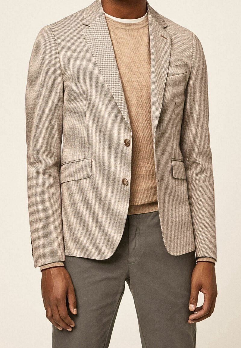 Beiger kariertes Sakko mit zwei Fronttaschen und Einzelknopfverschluss, kombiniert mit einem hellbeigen Pullover mit Rundhalsausschnitt und grauen Hosen.