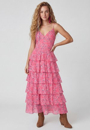 Jonge vrouw met lang krullend haar, gekleed in een mouwloze roze maxi-jurk met bloemenprint en laagjes, en bruine enkellaarzen, poserend met de hand in de zij.