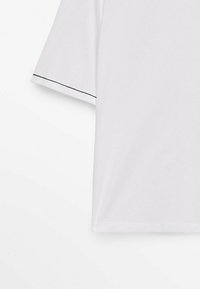 Chemise blanche à manches courtes avec un détail de surpiqûre noire le long du bord de la manche sur un fond blanc uni.