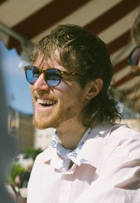 Γυαλιά ηλίου με σχέδιο χελώνας και μπλε φακούς, φορεμένα πάνω από ένα ανοιχτό ροζ πουκάμισο και ένα μπλε φουλάρι με σχέδια. Φυσικός φωτισμός σε εξωτερικό περιβάλλον.