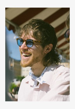 Teknőspáncél napszemüveg kék lencsékkel, világos rózsaszín ingre és kék mintás sálra viselve. Természetes fényben, szabadtéri háttérrel.