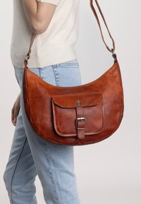 Kvinna iklädd ljusblå jeans och en krämfärgad tröja bär en stor brun lädercrossbody-väska med en framficka med spänne.